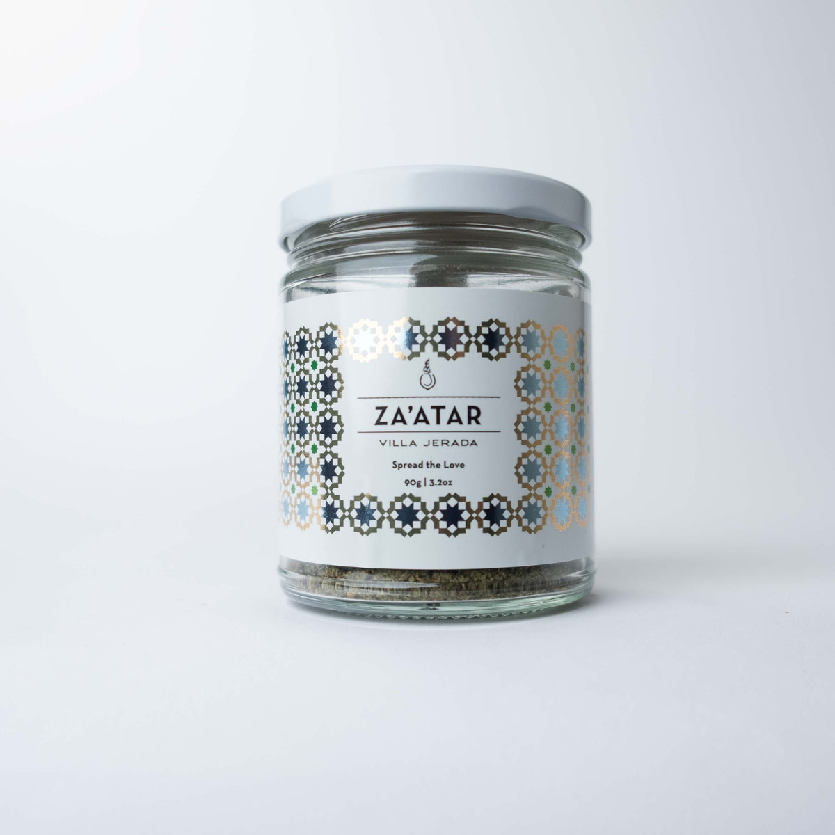 A clear glass jar filled with green-brown za'atar spice blend, featuring a minimalist white label and black text, resting on a clean white surface.