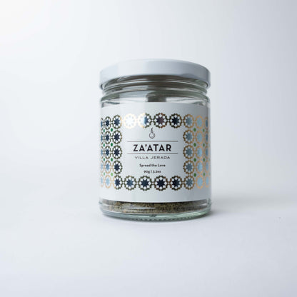 A clear glass jar filled with green-brown za'atar spice blend, featuring a minimalist white label and black text, resting on a clean white surface.
