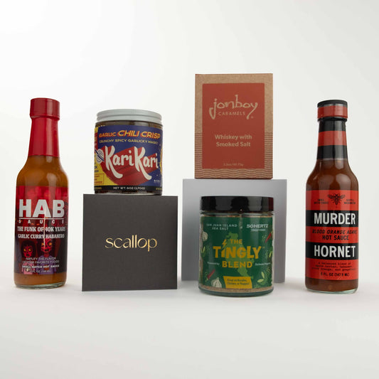 A flat lay of gourmet food products: HAB hot sauce, KariKari chili crisp, a black Scallop box, Jonboy caramels, Tingly Blend seasoning, and Murder Hornet hot sauce against a white background.