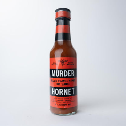 A bottle of Murder Hornet Blood Orange Agave Hot Sauce with a red label, black and white stripes, and a hornet illustration, set against a white background.